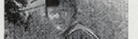 A grainy black and white photograph of Marion Lucy Queal in graduation regalia with trees and buildings in the background.