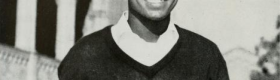 A black and white photograph of Sherrill Luke smiling, wearing a black sweater with a white college underneath, and a building with columns in the background.