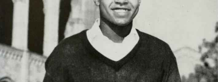 A black and white photograph of Sherrill Luke smiling, wearing a black sweater with a white college underneath, and a building with columns in the background.