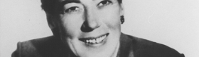 A black and white photograph of UCLA psychologist Evelyn Hooker smiling in a black coat against a white background.