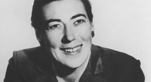 A black and white photograph of UCLA psychologist Evelyn Hooker smiling in a black coat against a white background.