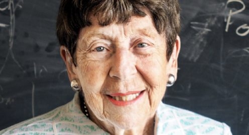 A head and shoulders photo of Assistant research geophysicist Margaret Kivelson in a white and green coat standing in front of a chalkboard.