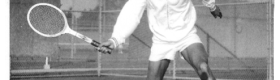 A black and white photograph depicting Arthur Ashe in white shorts and a white sweater on a tennis court, a tennis racket in his right hand.