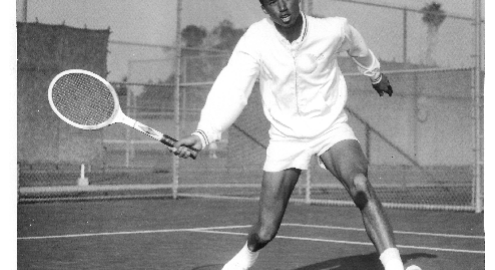 A black and white photograph depicting Arthur Ashe in white shorts and a white sweater on a tennis court, a tennis racket in his right hand.