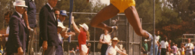 Jackie Joyner-Kersee in blue and gold UCLA exercise clothing performing a long distance jump with spectators in the background.