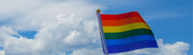 An image of someone holding a miniature rainbow flag or pride flag with clouds and a blue sky in the background.