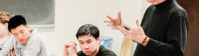 A male professor lecturing in a class room with a chalkboard with two male students listening attentively.