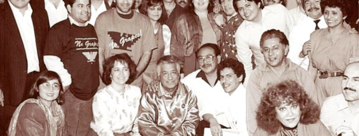 A black and white photograph of César E. Chávez surrounded twenty plus people as they pose for the photograph.