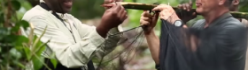 Two men in a jungle environment conducting an experiment involving a wooden branch with a sharp point and a thin black net.