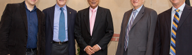 Physicist Mani Bhaumik (center) dressed in a black suit with a white shirt and pink tie, surrounded by two mean in suits to both his left and right, one of whom is Chancellor Block in a grey suit, standing in front of a building with gold letters spelling out, "Mani L. Bhaumik Institute for Theoretical Physics," which was created thanks to Mr. Bhaumik $11 million donation, the largest gift in the history of the UCLA Division of Physical Sciences.