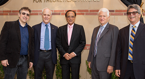 Physicist Mani Bhaumik (center) dressed in a black suit with a white shirt and pink tie, surrounded by two mean in suits to both his left and right, one of whom is Chancellor Block in a grey suit, standing in front of a building with gold letters spelling out, "Mani L. Bhaumik Institute for Theoretical Physics," which was created thanks to Mr. Bhaumik $11 million donation, the largest gift in the history of the UCLA Division of Physical Sciences.