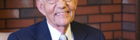 Alumnus Morton La Kretz wearing a blue tie with a white collar shirt and light blue tie, with a red brick wall in the background.