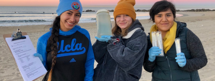 Three females students with lab equipment standing by a sandy beach, with waves and a multicolored sky in the background.