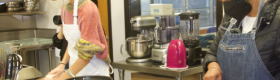 Two female students wearing masks in an indoor kitchen setting slicing apples and other food items.