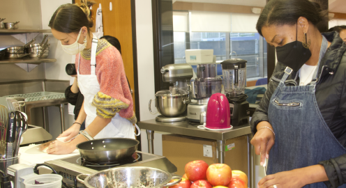 Two female students wearing masks in an indoor kitchen setting slicing apples and other food items.