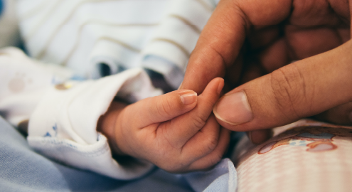 An adult's hand gently grabbing the hand of a newborn baby which is wrapped in a white pajama and cradled by blankets.