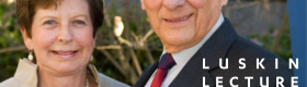 Meyer Luskin wearing a blue suit with a red tie and Renee Luskin in a beige dress with a pearl necklace standing outside with tree branches and pink and white flowers in the background.