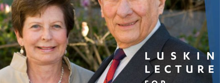 Meyer Luskin wearing a blue suit with a red tie and Renee Luskin in a beige dress with a pearl necklace standing outside with tree branches and pink and white flowers in the background.