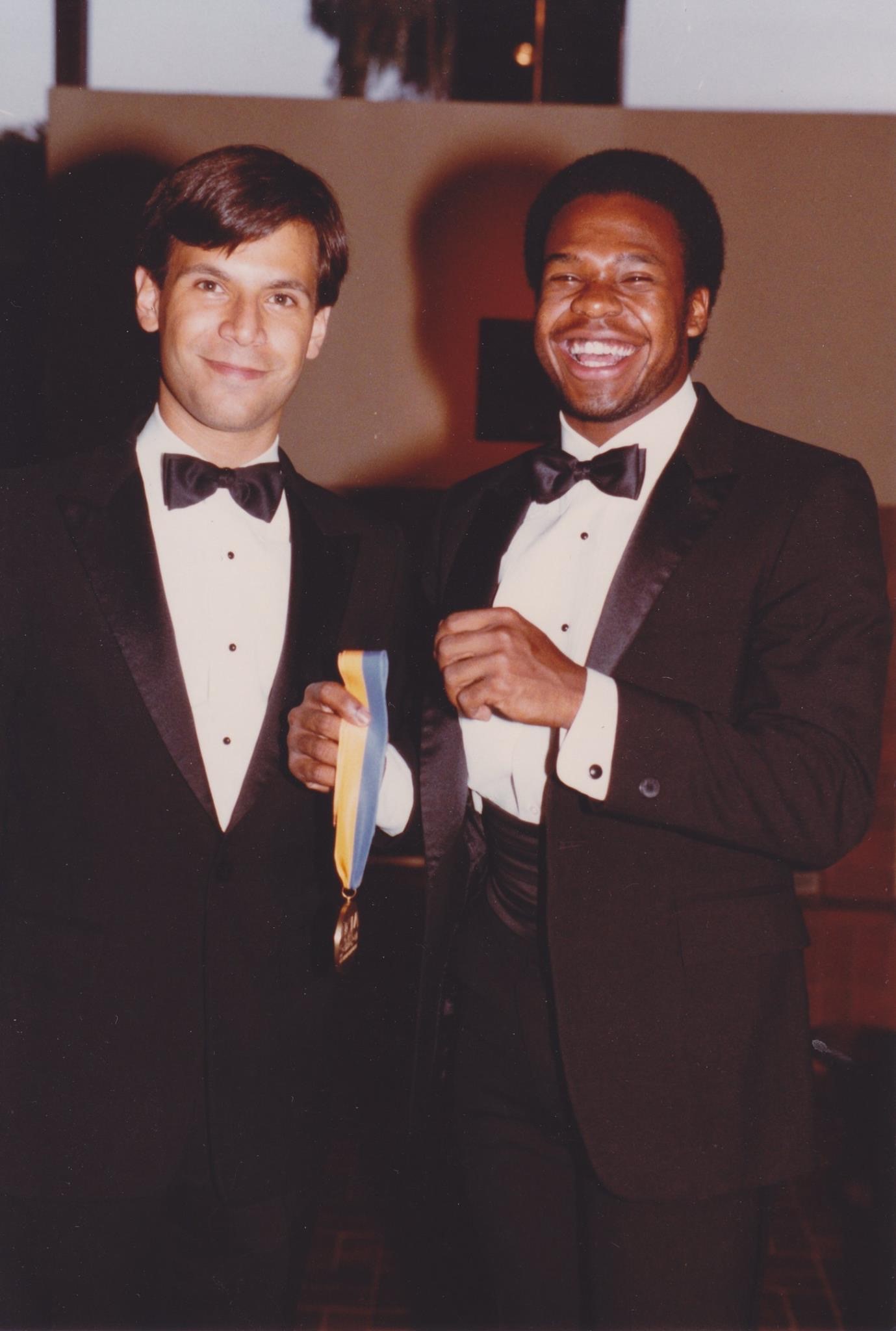 A photograph of Robert "Bobby" Grace in a tuxedo holding a gold medal with a blue and yellow ribbon standing next to another man, also wearing a tuxedo.