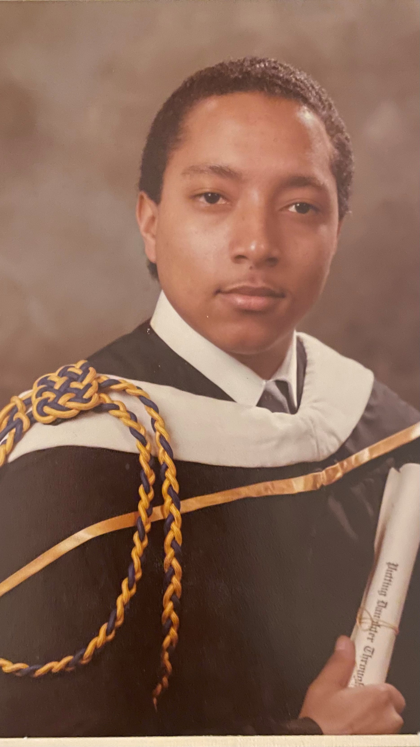 A photograph of Brett York wearing graduation regalia holding a diploma with a blue and yellow graduation cord hanging from one of his shoulders.