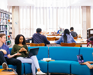 Student sitting inside a well lit room with a blue couch and communal desks in the background.