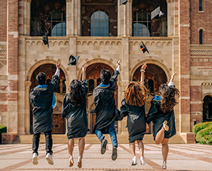 A group of jubilant graduates in traditional black caps and gowns are captured mid-jump against a backdrop of Royce Hall with it's famed arched entryways. The graduates, facing away from the camera, are seen tossing their caps high into the air, marking a celebratory moment under the clear, sunny skies that signify the bright future ahead.