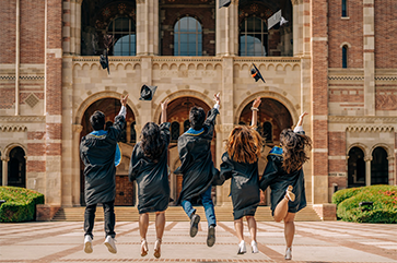 A group of jubilant graduates in traditional black caps and gowns are captured mid-jump against a backdrop of Royce Hall with it's famed arched entryways. The graduates, facing away from the camera, are seen tossing their caps high into the air, marking a celebratory moment under the clear, sunny skies that signify the bright future ahead.