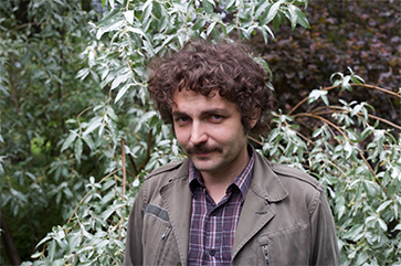 Boris Dralyuk standing in front of green foliage while wearing a green jacket over a plaid shirt.