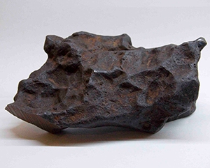 A meteorite fragment against a white background with an out-of-view overhead light source creating a shadow as the base of it.