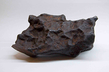 A meteorite fragment against a white background with an out-of-view overhead light source creating a shadow as the base of it.