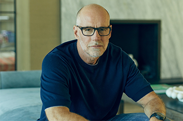 Scott Galloway wearing a blue t-shirt, sitting down in an indoor setting with a fire place in the background.