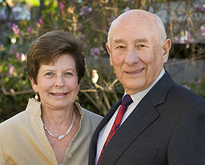 Renee (left) and Meyer Luskin standing outside on a sunny day with green leaves, branches and pink flowers in the background.
