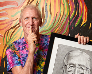 Bill Walton wearing a tie die shirt with his right index finger pressed to this lips while holding a black and white drawing of John Wooden against a painted wall with orange, yellow, red, green, brown and black streaks of paint.