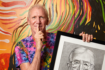 Bill Walton wearing a tie die shirt with his right index finger pressed to this lips while holding a black and white drawing of John Wooden against a painted wall with orange, yellow, red, green, brown and black streaks of paint.