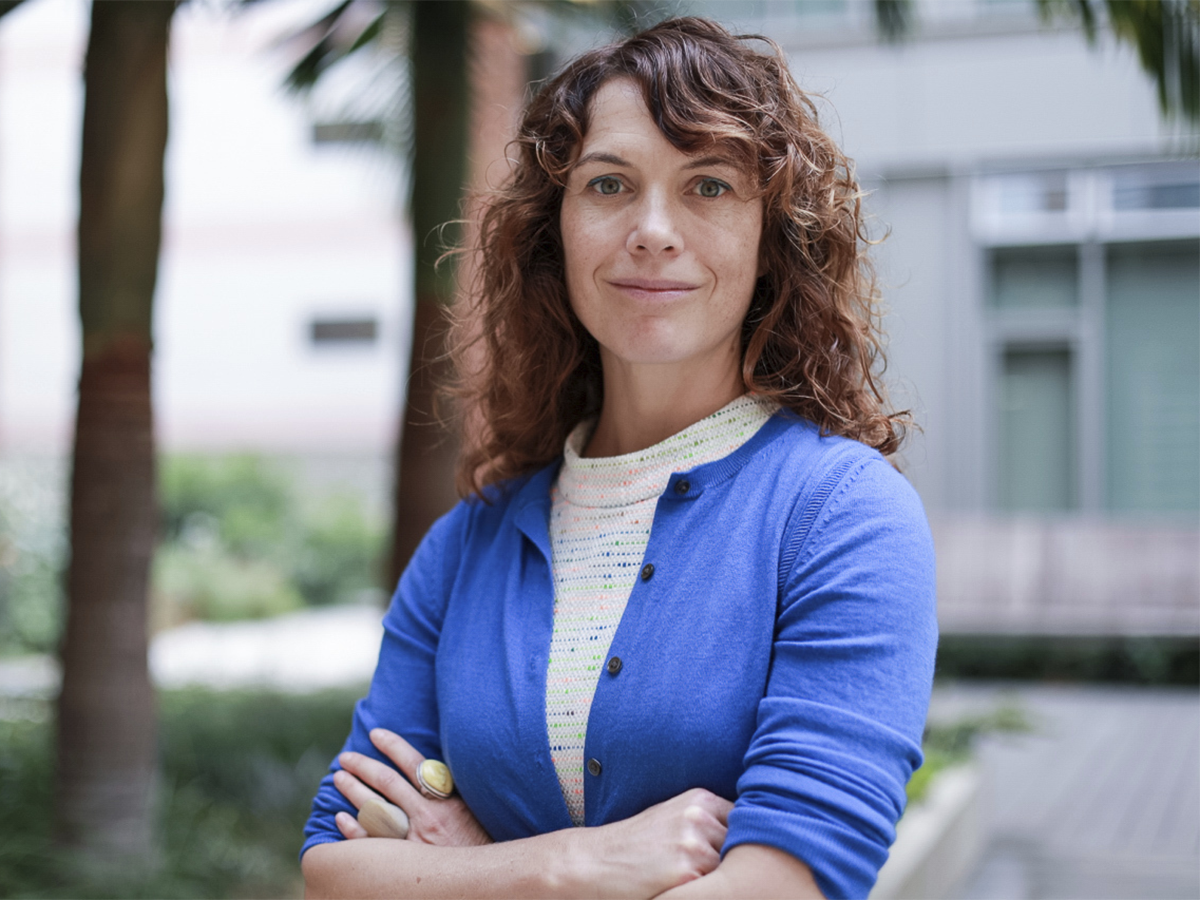 Amy Rowat in a blue sweater with arms crossed against a blurred outdoor background with palms trees. 