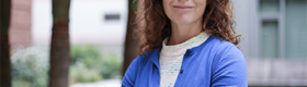 Amy Rowat in a blue sweater with arms crossed against a blurred outdoor background with palms trees.