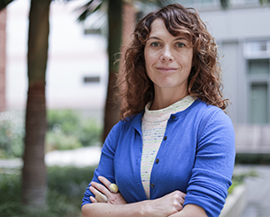 Amy Rowat in a blue sweater with arms crossed against a blurred outdoor background with palms trees.