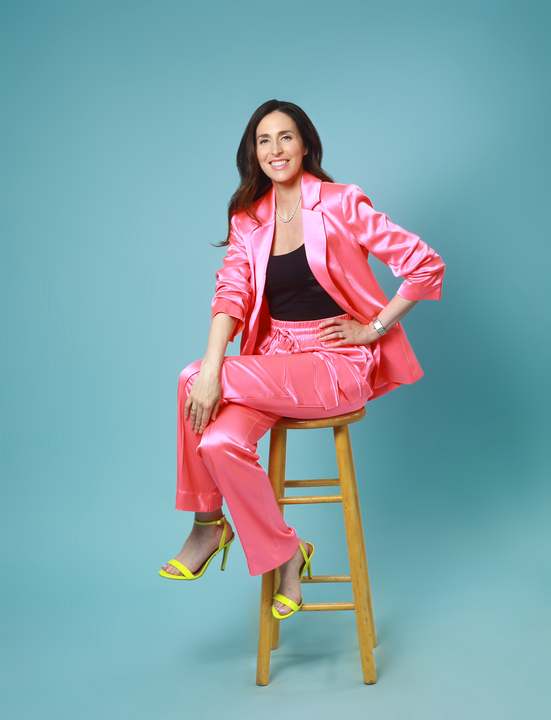 Hannah Gordon is a pink suit with a black top, sitting on a wooden barstool in front of a light blue photo studio background. 