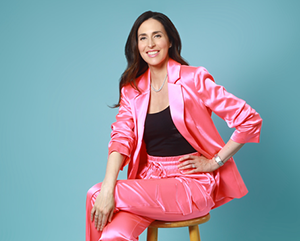 Hannah Gordon is a pink suit with a black top, sitting on a wooden barstool in front of a light blue photo studio background.