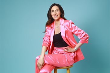 Hannah Gordon is a pink suit with a black top, sitting on a wooden barstool in front of a light blue photo studio background.