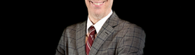 Jack Bobo in a grey suit and red tie against a black background.