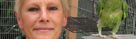 Ursula Heise inside a cage with a green parrot perched on a ledge close to her face.