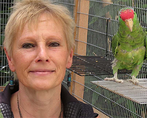 Ursula Heise inside a cage with a green parrot perched on a ledge close to her face.