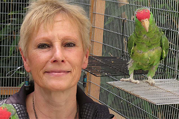 Ursula Heise inside a cage with a green parrot perched on a ledge close to her face.