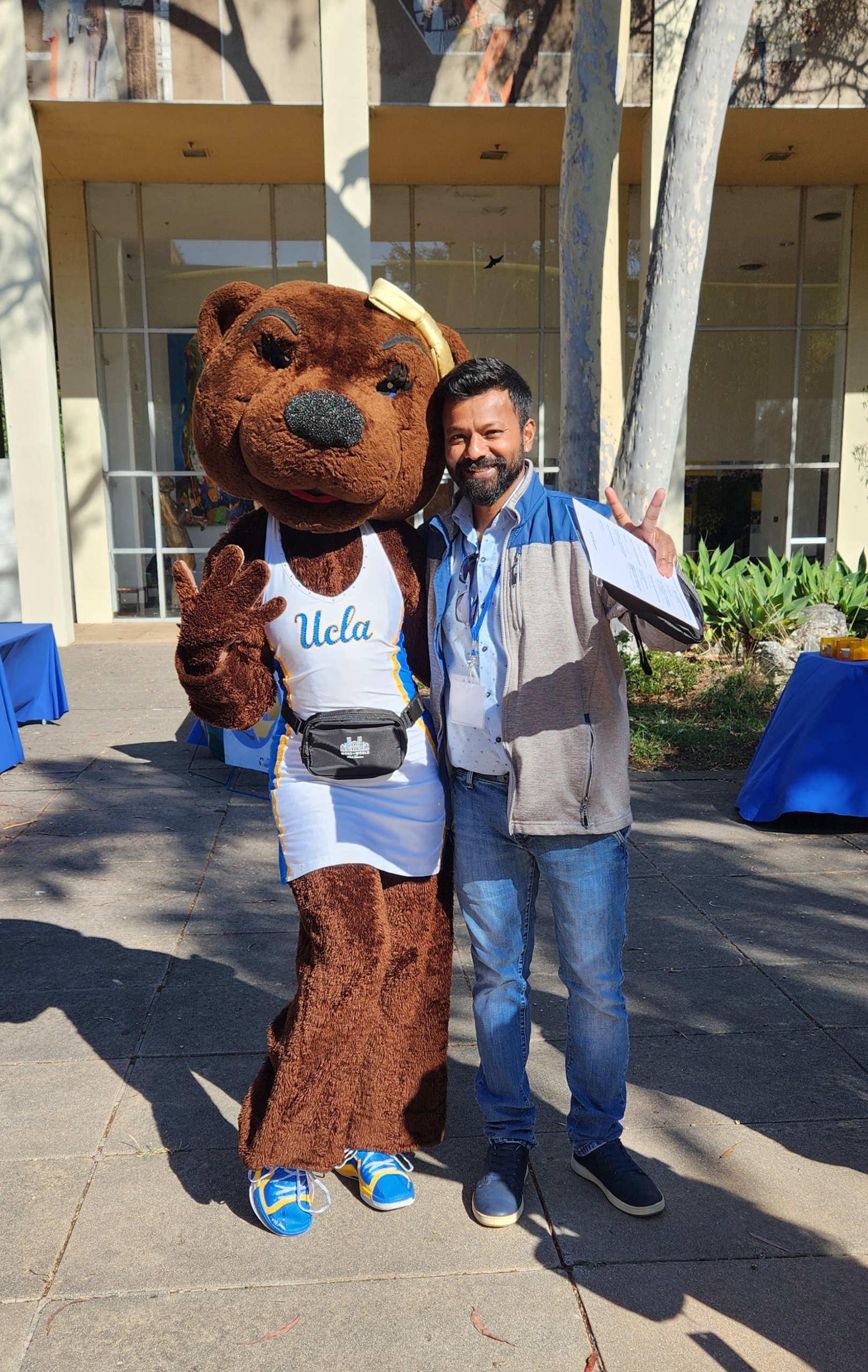 Vetri Nathan poses on the UCLA campus while wearing a blue and gray jacket, standing next to Josie Bruin the mascot.