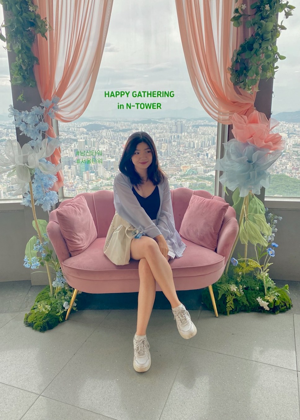 Claudia Chen sitting on a pink couch in front of a window with a view of Seoul, framed with flowers, greenery and peach curtains.