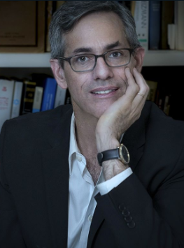 David Myers with his left hand cupping his chin; he wears glasses, a dark suit and a black and gold watch.