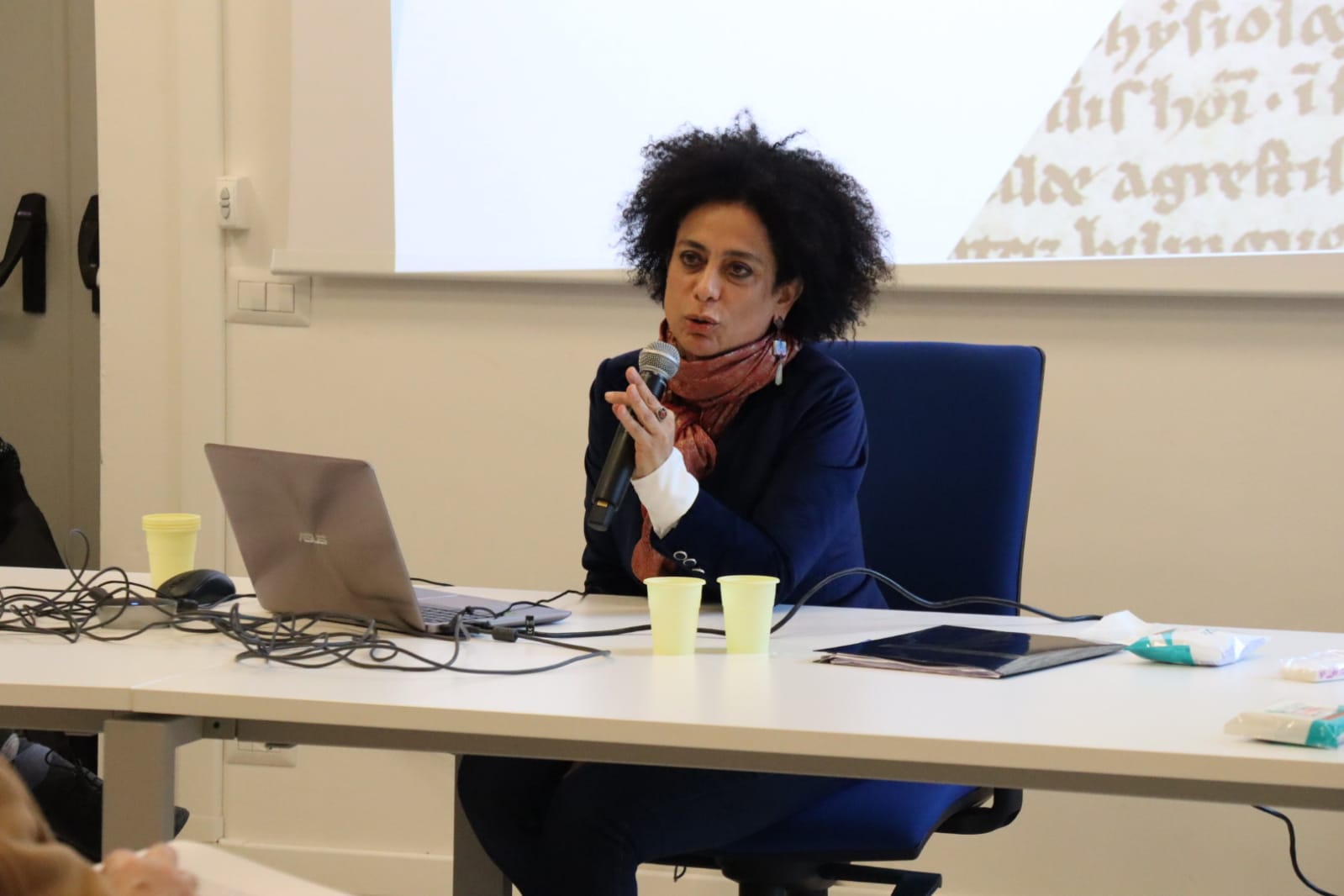 Roberta Morosini in a blue suit with a rust-colored scarf holds a microphone and delivers a presentation while seated at a table.