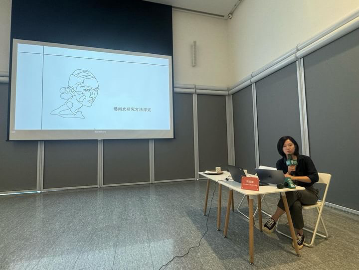 A wide shot of a classroom with a screen and a presentation projected on it; Pin-Hua Chou sits at a table in the lower right of the picture holding a microphone.