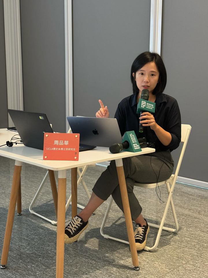 Pin-Hua Chou seated at a table, holding a microphone and delivering a presentation